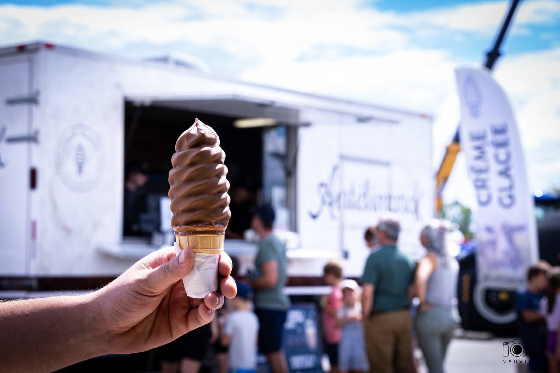 Une main tenant un cornet de crème glacée enrobée de chocolat devant la crèmerie mobile d'Antélimarck.