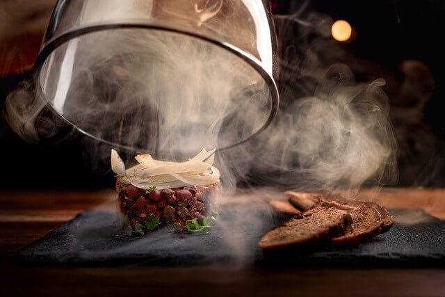 Une main soulevant une cloche de verre pour dévoiler un tartare du Pub L'Ours Noir enveloppé de fumée.